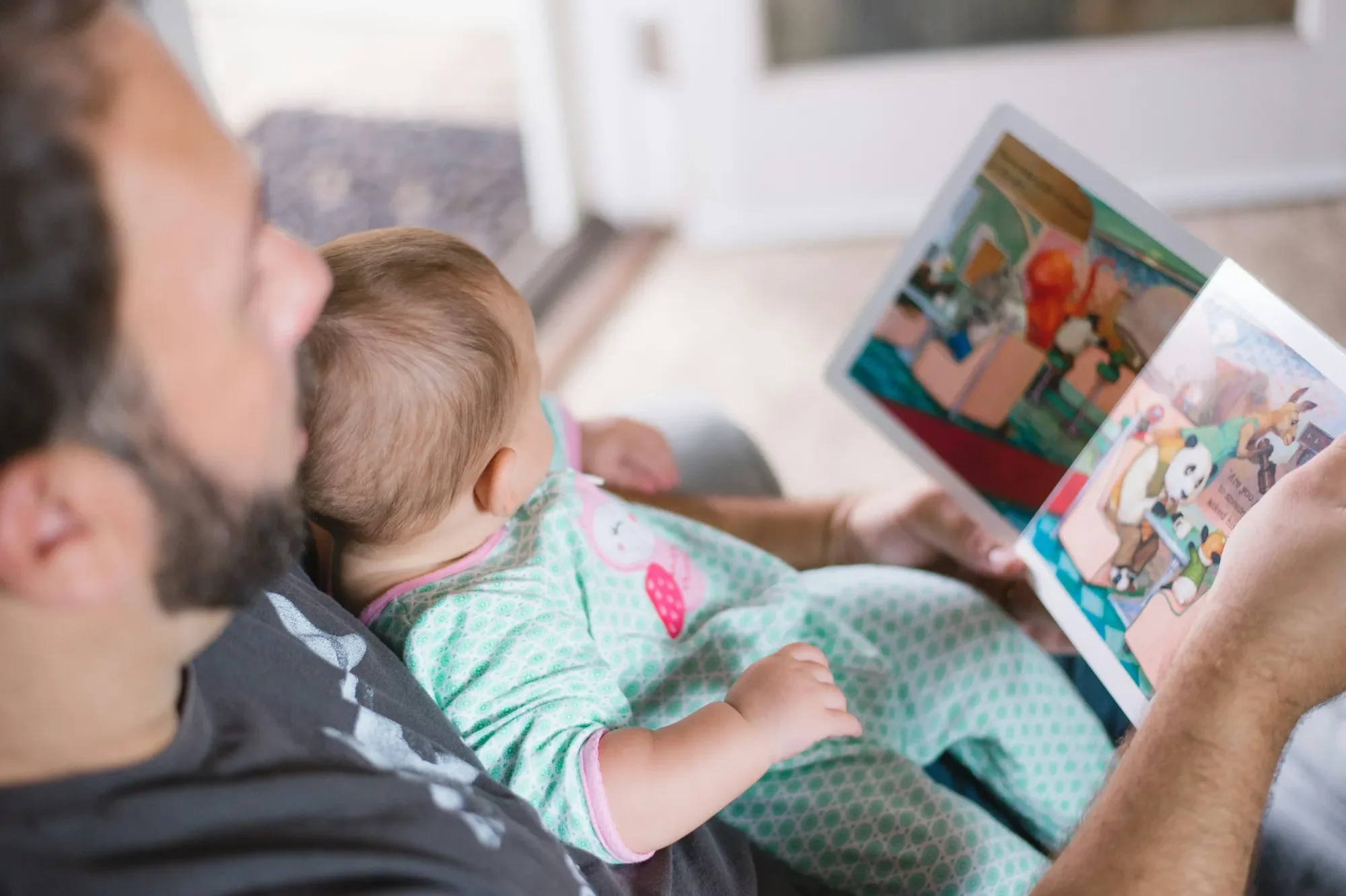 Sozial-Emotionales Lernen Vater liest einem Baby ein Bilderbuch vor, beide entspannt im Wohnraum.